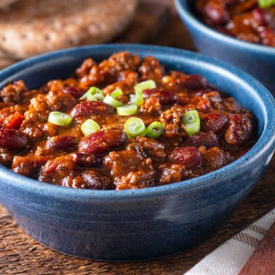 Beef chili con carne topped with green onions in blue bowl on wood counter