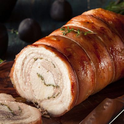 Homemade sliced porchetta roll topped with rosemary on wood board beside knife