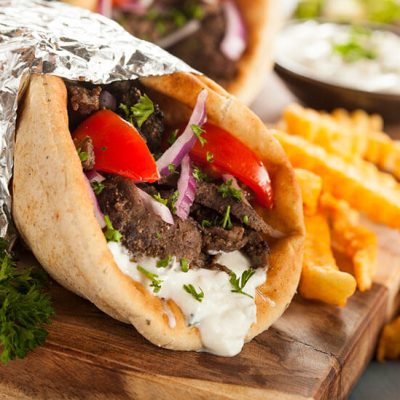 Beef shawarma wrapped in foil with tzatziki sauce on wood board beside fries